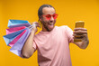 © wpadington - Studio shot of young African American man customer holding a bunch of shopping bags and looking at his smartphone screen with excited open mouth face expression, isolated on yellow background