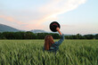 © Elsa - Sunset picture of a woman in a green wheat field holding a black hat in her hand. Peace of mind, freedom, and a deep connection with nature. Pastel sky, clouds, mountains, and forest in the background