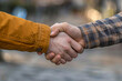 © EliteStock - Two hands, one wearing an orange jacket and the other in a plaid shirt, engaged in a firm handshake symbolizing unity and partnership.