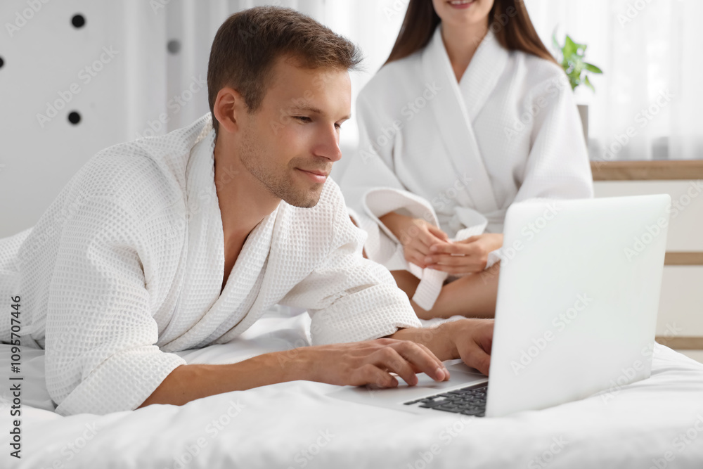 Young man in bathrobe using laptop with his wife on bed at home