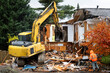 © knelson20 - Large yellow excavator starting demolition on a house, workman spraying water to keep dust down, rainy day residential construction project job site