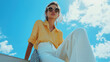 © ibooM - A woman poses confidently in a yellow shirt and white pants under a bright blue sky