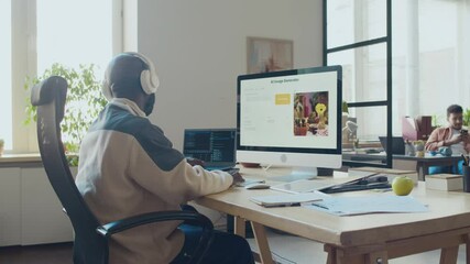 Medium full side shot of African American man in headphones sitting at desk at IT company, typing computer code while working on image generation software, in cozy office at daytime