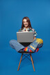 © Davidovici - Vertical photo of smiling young student girl in shirt and headphones working on laptop while sitting on chair with crossed legs, isolated on blue background.