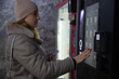© Андрей Рыков - Side view portrait of adult caucasian woman in winter clothes (grey jacket and hat) choosing hot drink in black free standing bean-to-cup coffee vending machine. Soft focus. Food business theme.
