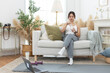 © eakgrungenerd - Asian young female sporty holding water bottle sitting on sofa in living room while exercising. stay hydrated per day. Healthy fit woman fresh wellness. Drinking water makes you feel good.
