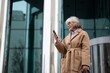 © Ivan Traimak - Senior woman in stylish beige coat checks her smartphone outside a modern building on a clear day