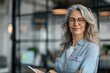 © btiger - Confident middle-aged business woman standing in office with tablet, smiling senior female executive wearing glasses and holding digital pad near glass wall, professional corporate portrait