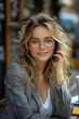 © Gonzalo - Cheerful young woman with blonde wavy hair and glasses, speaking on the phone while working on a laptop outdoors, bathed in natural sunlight with a relaxed and professional demeanor.