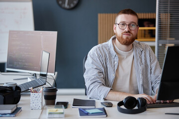 Portrait of bearded man looking at camera while sitting at desk in modern office setup with various gadgets and tools around him