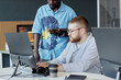© Seventyfour - Two men working together at desk with computer equipment, discussing tasks on digital devices, in professional setting promoting teamwork and efficiency