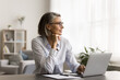 © fizkes - Happy dreamy mature freelancer woman in stylish eyeglasses working at home office table, sitting at laptop computer, looking away, smiling, touching chin, thinking, using pc for online communication,