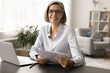 © fizkes - Happy beautiful grey haired mature business woman in elegant eyeglasses sitting at home office workplace, holding paper document at laptop, looking for portrait at camera, smiling
