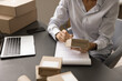 © fizkes - Cropped shot of small webstore owner processing order for customer, writing notes in register at workplace, holding small paper wrap, package with purchase for shipping