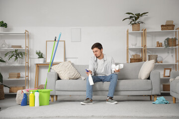 Poster - Stressed young man with cleaning supplies sitting on sofa at home