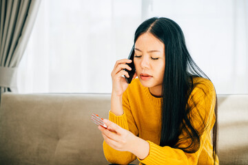 An unwell young Asian woman sits on a couch holding medicine using her phone to consult a doctor at home. Depicting medication intake medical advice and quarantine during social distancing.