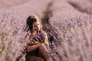 Wall Mural - Girl is sitting in a field of purple flowers. She is holding a basket of flowers and smiling. Scene is peaceful and serene, as the girl is surrounded by the beauty of nature.