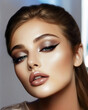 © stefanholm - A glamorous close-up portrait of a young Caucasian woman with shimmering champagne eyeshadow, precise eyeliner, and glossy nude lips, glowing under soft studio lighting.