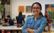 © SOMMAI - A smiling businesswoman stands with her arms crossed in an office, with a team working in the blurred background.