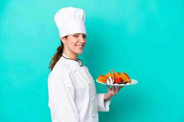 Wall Mural - Young caucasian chef woman holding waffles isolated on blue background smiling a lot