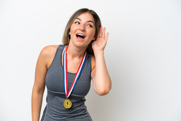 Young Rumanian woman with medals isolated on white background listening to something by putting hand on the ear