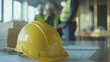 © Prapan - a bright yellow safety helmet placed on a modern office desk, with blueprints and project plans scattered around. In the background,