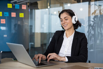 Woman in business suit smiling, typing on laptop at office desk with headphones. Engaged in online work, multitasking in modern workplace.