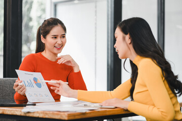 Wall Mural - Two women discussing business report with charts and graphs, showcasing teamwork and collaboration in modern office setting