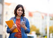 © Prostock-studio - Smiling Young Middle Eastern Female Student Walking With Smartphone And Workbooks On City Street, Happy Millennial Arab Woman Messaging On Mobile Phone While Going Home After Classes, Copy Space