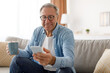 © Prostock-studio - Portrait of smiling mature man in eyeglasses using mobile phone, watching video and holding cup of coffee, sitting on the couch in living room. Guy browsing internet, surfing web, free copy space