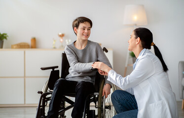 Disabled people healthcare support concept. Doctor holding hands of handicapped teen boy in wheelchair during medical visit. Happy impaired adolescent and young nurse at home