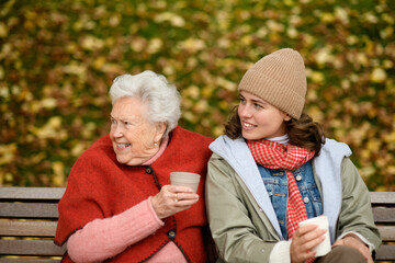 Wall Mural - Portrait of a granddaughter and grandmother sitting on bench in autumn park, drinking coffee and laughing together.