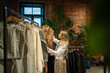 © DimaBerlin - Interested woman stands in fashion store, carefully chooses clothes to buy. Pleased female looks at stylish dress while shopping, holds hanger with item, tries on likes, examining before make decision