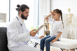 © Sofiia Shunkina - Caucasian male doctor checks little girl with headache at medical clinic. Young patient shows discomfort while doctor reviews information on tablet in professional environment.