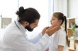 © sofiko14 - Caucasian male doctor examines throat of sick girl in medical office for enlarged lymph nodes. Medical professional checks young patient during routine health examination focusing on wellness and care