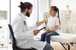 © sofiko14 - Male doctor talks with young white girl sitting on medical couch, recording details in tablet. Scene in bright examination room with relaxed and comfortable atmosphere.