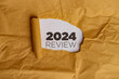 © Jamaludinyusup - A yellow piece of paper with a hole torn in it. Through the hole, a white piece of paper is visible with the text '2024 REVIEW' written in black. This represents the concept of reviewing the year 2024