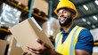 © LifeMedia - A young male worker wearing a hard hat and reflective vest smiles as he holds a cardboard package in the warehouse aisle, surrounded by high shelves of goods.