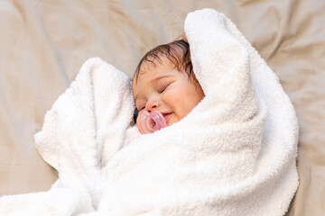 Wall Mural - A beautiful newborn baby girl lying on bed wrapped in white terry towel after bathing and smiling. baby hygiene concept. Top view. High quality photo