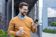 © Tetiana - A young smiling man is standing outside an office building, holding a cup of coffee and using a mobile phone
