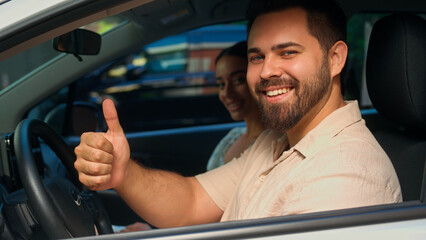 Happy smiling Caucasian male guy driver car owner buyer sitting inside new auto with woman female girl passenger man client looking camera showing thumb up recommend family buying vehicle automobile