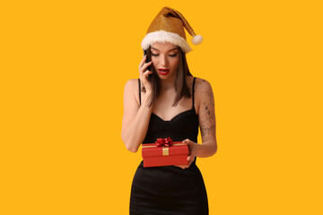 Wall Mural - Young woman in Santa hat talking by mobile phone and holding Christmas gift box on yellow background