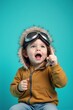 © Alex - Joyful toddler in aviator goggles and furry hat pointing upwards against a bright blue background