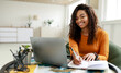 © Prostock-studio - Smiling young African American woman sitting at desk working on laptop taking notes in notebook, happy millennial female studying online, watching webinar using computer and writing check list