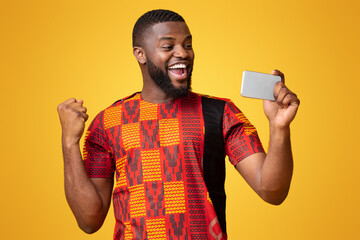 Wall Mural - Happy black man in african t-shirt holding phone and expressing happiness, shouting and clenching fist, yellow background