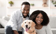 © Prostock-studio - Love And Care Concept. Portrait of cheerful black couple patting their happy labrador spending time together in living room at home, embracing golden retriever pet, posing looking at camera