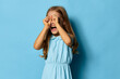 © SHOTPRIME STUDIO - Little girl in a blue dress covering her eyes with her hands, standing against a matching blue background