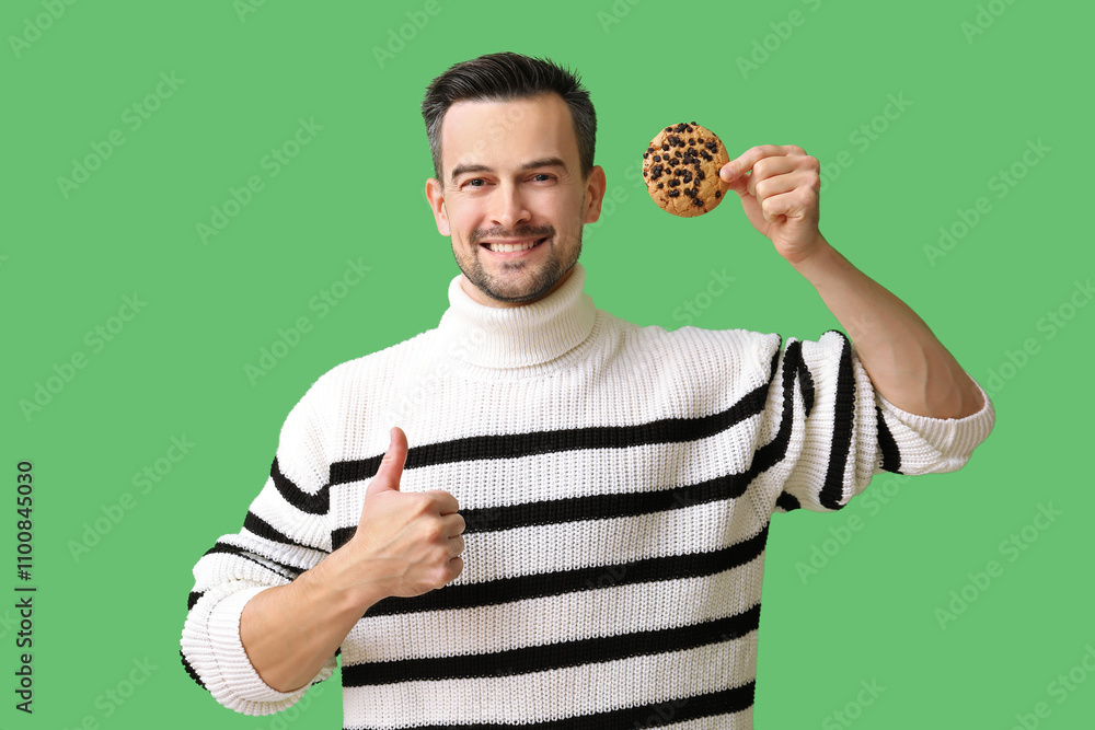 Handsome man with tasty cookie showing thumb-up on green background