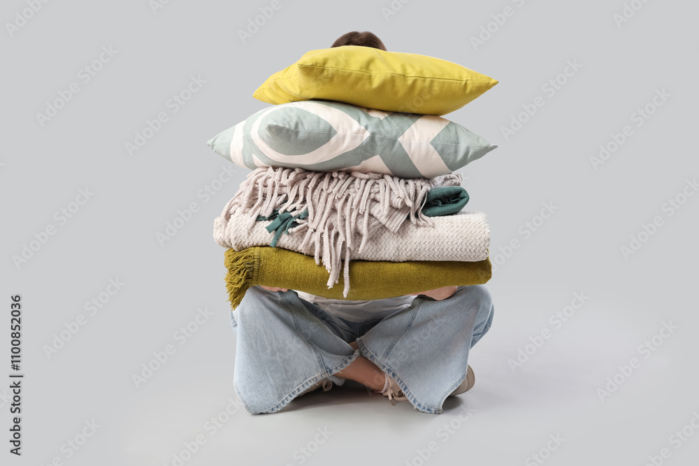 Beautiful young woman with stack of different blankets and pillows sitting on grey background