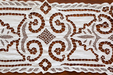 Fragment of handmade tablecloth with lace patterns embroidered in richelieu style or cutwork embroidery on wooden table. Vintage doily texture. Retro handicraft for home decoration. Flat lay, close-up